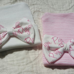 Op de afbeelding: Twee baby mutsen, een witte en een roze gestreepte, elk versierd met een witte strik. De strikken zijn bedrukt met de naam "Piper" in roze script. De mutsen zijn gemaakt van een zacht, geribbeld materiaal.