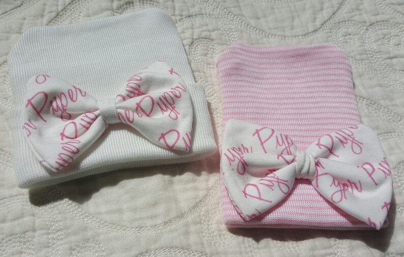 Op de afbeelding: Twee baby mutsen, een witte en een roze gestreepte, elk versierd met een witte strik. De strikken zijn bedrukt met de naam "Piper" in roze script. De mutsen zijn gemaakt van een zacht, geribbeld materiaal.