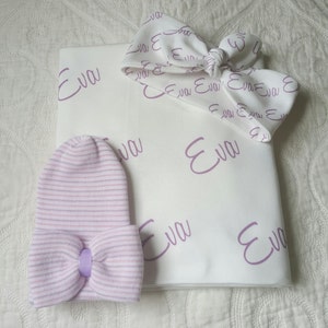 May include: A white baby blanket with the name "Eva" printed in purple script. A matching purple and white striped hat with a bow is included. A purple headband with a bow is also included.