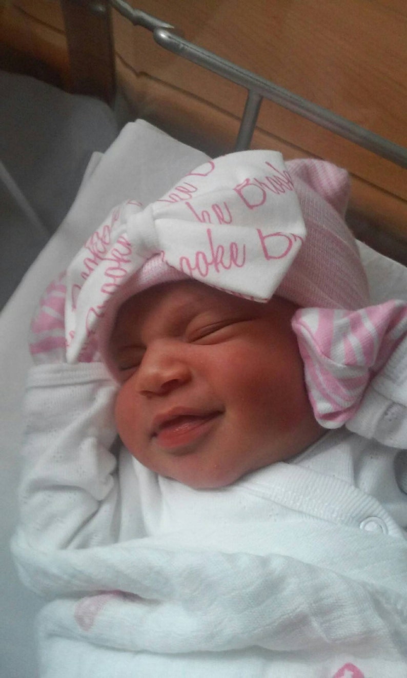 Op de afbeelding: Een pasgeboren baby met een roze en witte gestreepte hoed met een strik en de naam "Brooke" erop gedrukt. De baby draagt een witte onesie en glimlacht met gesloten ogen.