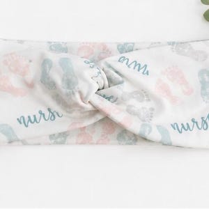 Pu&ograve; includere: Fascia bianca con un design a nodo a torsione. La fascia presenta impronte di piedi di bambino in rosa, grigio e blu, con la parola "nurse" in blu. Un rametto di verde &egrave; sullo sfondo.