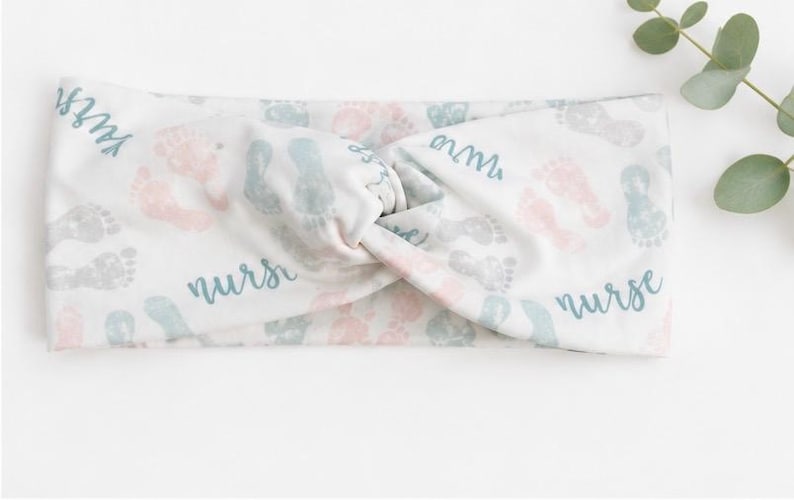Pu&ograve; includere: Fascia bianca con un design a nodo a torsione. La fascia presenta impronte di piedi di bambino in rosa, grigio e blu, con la parola "nurse" in blu. Un rametto di verde &egrave; sullo sfondo.