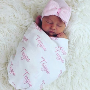 May include: A white swaddle blanket with the name "Teagan" printed in pink script all over. A baby is swaddled in the blanket and is wearing a pink and white striped hat with a bow.