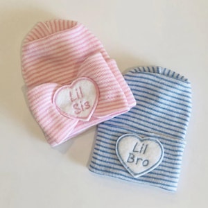 May include: Two striped baby hats, one pink and one blue. Each hat has a white heart-shaped patch with embroidered text. The pink hat says "Lil Sis," and the blue hat says "Lil Bro." These are newborn baby hats.
