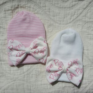 Op de afbeelding: Twee baby mutsen. E&eacute;n is roze en wit gestreept, de andere is wit. Beide hebben een witte strik met de naam "Piper" in roze script. De mutsen zijn gemaakt van zachte gebreide stof.