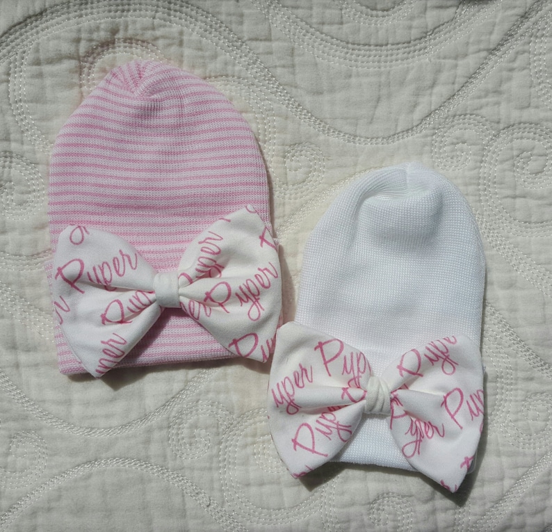 Op de afbeelding: Twee baby mutsen. E&eacute;n is roze en wit gestreept, de andere is wit. Beide hebben een witte strik met de naam "Piper" in roze script. De mutsen zijn gemaakt van zachte gebreide stof.
