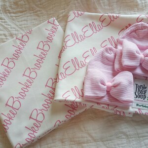 May include: Two white baby blankets with pink personalised lettering, one with the name "Ella" and the other with the name "Brooke". Two pink striped baby hats with bows are included, along with a small white card with the text "Turtle Bug Soup #turtlebugsoup".