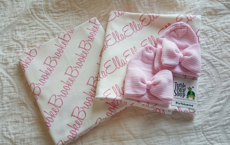 May include: Two white baby blankets with pink personalised lettering, one with the name "Ella" and the other with the name "Brooke". Two pink striped baby hats with bows are included, along with a small white card with the text "Turtle Bug Soup #turtlebugsoup".