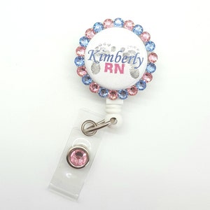 May include: A white badge reel with a pink and blue rhinestone border. The badge has a silver background with the text "Kimberly RN" and a pair of baby footprints. The reel has a clear plastic strap with a pink rhinestone.
