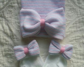 May include: A set of baby accessories in pink and blue. The set includes a striped knit hat with a matching bow, and two pairs of white socks with striped bows.