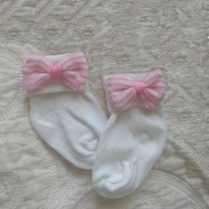May include: A pair of white baby socks with pink and white striped bows on the top of each sock.