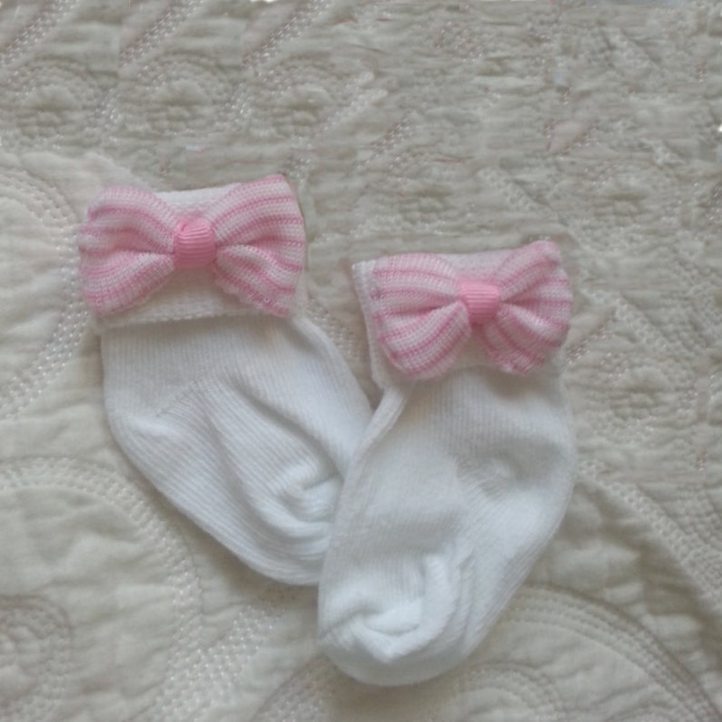 May include: A pair of white baby socks with pink and white striped bows on the top of each sock.