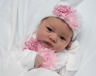 May include: A newborn baby wearing a white onesie with pink and white flower embellishments and a white hat with pink and white flowers.