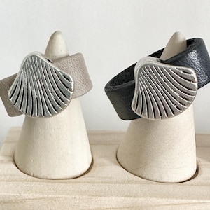 Könnte beinhalten: Zwei Lederringe mit silbernen Muscheldesign werden auf weißen, kegelförmigen Ständern präsentiert. Ein Ring hat ein graues Lederband, der andere ein schwarzes Lederband. Die Ringe befinden sich auf einem hellen Holztablett.