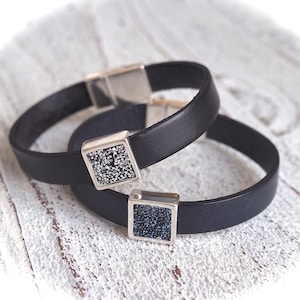 Könnte beinhalten: Zwei schwarze Lederarmbänder mit silbernen quadratischen Verschlüssen. Ein Verschluss ist mit klaren Strasssteinen besetzt, der andere mit blauen Strasssteinen.