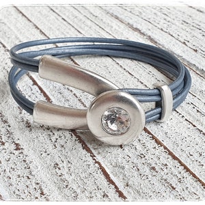 Könnte beinhalten: Ein graues Lederarmband mit einem silbernen Verschluss und einem klaren Strassstein. Das Armband besteht aus mehreren Ledersträngen und hat ein einzigartiges Design.