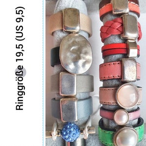 Könnte beinhalten: Ein Set aus neun silbernen Ringen mit verschiedenen Designs, darunter ein runder Ring mit strukturierter Oberfläche, ein rechteckiger Ring und ein Ring mit einem kleinen blauen Stein. Die Ringe befinden sich auf einem Präsentationsständer mit weißem Hintergrund. Ringgröße 19,5 (EU 9,5).