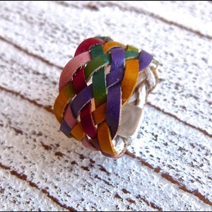 May include: A multi-coloured leather ring with a braided design in shades of pink, red, green, purple, and yellow. The ring is made of woven leather strips and has a silver metal base.