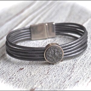 Könnte beinhalten: Ein graues Lederarmband mit einem Magnetverschluss und einem runden, silberfarbenen Anhänger mit einer dunkelgrauen, gesprenkelten Oberfläche.
