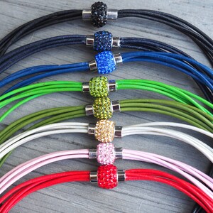 Pu&ograve; includere: Un set di otto collane in cordoncino di pelle con perline di cristallo di vari colori. Le collane sono disposte a forma di arcobaleno, con nero, blu, verde, oliva, bianco, rosa, rosso e nero.