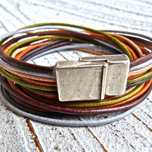 Könnte beinhalten: Mehrfarbiges Lederarmband mit einem silbernen Verschluss. Das Armband besteht aus mehreren dünnen Ledersträngen in den Farben Braun, Orange, Grün und Grau.