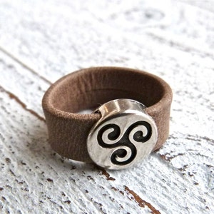 Könnte beinhalten: Ein brauner Lederring mit einem silbernen Metallaccent, der ein stilisiertes dreifaches Spiralmuster zeigt.