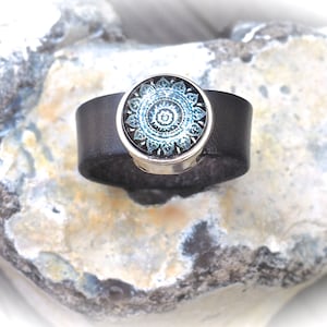 May include: A black leather bracelet with a silver-toned round charm featuring a blue and white mandala design.