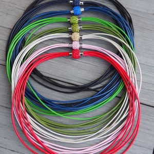 Pu&ograve; includere: Un set di otto collane in cordoncino di pelle con una variet&agrave; di perline di cristallo colorate. Le collane sono disposte in cerchio, con le perline rivolte verso l'esterno. I colori delle perline sono nero, blu, verde, oro, rosa e rosso.