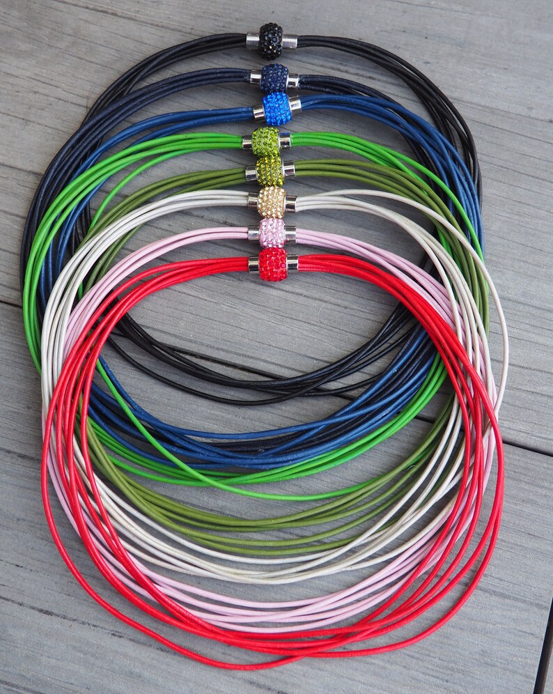 Pu&ograve; includere: Un set di otto collane in cordoncino di pelle con una variet&agrave; di perline di cristallo colorate. Le collane sono disposte in cerchio, con le perline rivolte verso l'esterno. I colori delle perline sono nero, blu, verde, oro, rosa e rosso.