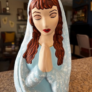 May include: A ceramic statue of a woman with closed eyes, wearing a light blue veil and robe. Her hair is brown, and her lips are red. Her hands are clasped in prayer. The statue has a serene expression.