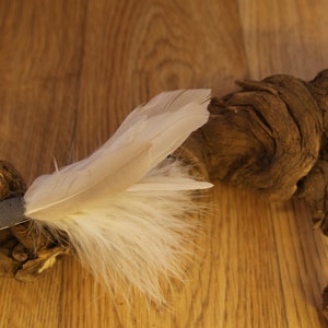May include: A decorative hair clip featuring a cluster of white feathers attached to a small, gray fabric base. The clip is set against a backdrop of a textured, dark brown wooden branch and a light brown wooden surface.