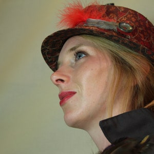 Peut inclure: Chapeau à motifs rouges et bruns avec une plume rouge et un bouton décoratif. Le chapeau est porté par une personne aux cheveux blonds et au rouge à lèvres rouge. Le chapeau semble être de style vintage ou steampunk.