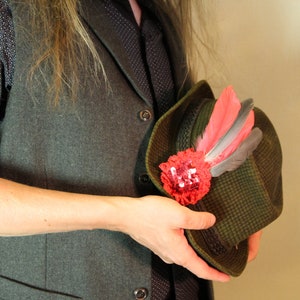 May include: A green hat with a decorative red flower and feathers. The hat has a patterned design and is held by a person wearing a gray vest and patterned shirt. The hat is a traditional style.