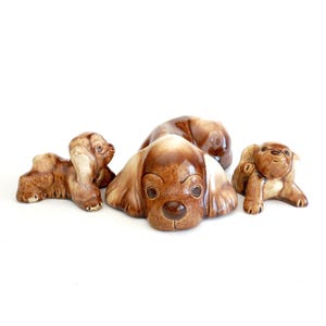 May include: Set of three ceramic dog figurines. The largest figurine is brown and white with a playful expression. The two smaller figurines are brown and white and are in a sitting position.