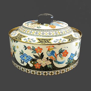 May include: A round, vintage tin container with a lid, featuring a colorful bird and floral design. The container is primarily white with black, gold, blue, orange, and yellow accents. The lid has a black knob.