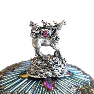 May include: A silver-toned figurine of a mythical creature with a pink gemstone on its back. The figurine is perched atop a decorative base with a textured surface.