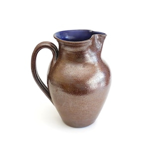 May include: A brown ceramic pitcher with a dark blue interior. The pitcher has a curved handle and a slightly flared spout. The exterior has a textured, speckled appearance, and the overall shape is rounded.