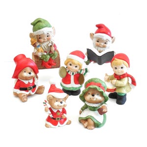 May include: A collection of seven ceramic figurines featuring various characters in holiday attire. The set includes elves, bears, and children wearing red and green hats, scarves, and outfits, with some holding items like a book, drum, and candy cane.