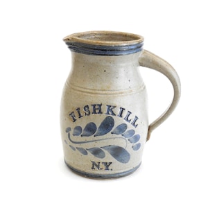 May include: A beige stoneware pitcher with a speckled finish, featuring a blue painted leaf motif and the text FISHKILL N.Y. in blue lettering. The vessel has a curved handle, a wide rim, and a blue band around the top and bottom edges.