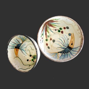 May include: Two round ceramic bowls with a cream-colored base and dark brown rims. Each bowl features a hand-painted design of a stylized yellow and blue creature with long, flowing fins, accented by small green and brown circular patterns.