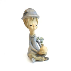 May include: A ceramic figurine of a seated figure wearing a blue hat and matching blue outfit. The figure has a pale complexion, closed eyes, and is holding a small, light-colored vase containing tiny blue flowers in their lap.