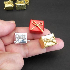 May include: Miniature gift boxes in red, silver, and gold, each with a decorative bow. The boxes are small, square-shaped, and appear to be wrapped. The image is a close-up, with the boxes held in a person's hand.