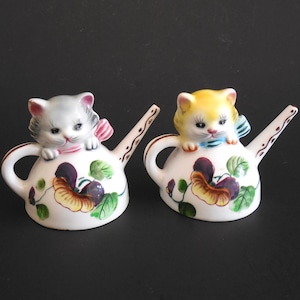 May include: Two ceramic cat figurines perched on white watering cans. One cat is gray with a pink bow, the other is yellow with a blue bow. The watering cans are decorated with floral patterns in shades of purple, yellow, and green.
