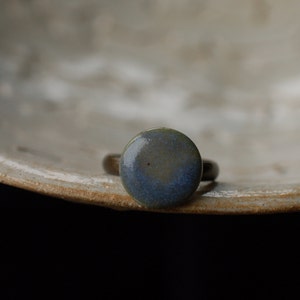 Könnte beinhalten: Ein handgefertigter Ring mit einem runden, glasierten Keramikstein in Blau- und Grüntönen. Der Ring hat ein dunkles Metallband. Der Ring wird auf einer strukturierten, cremefarbenen Oberfläche präsentiert.