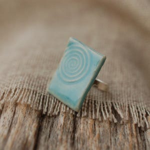Könnte beinhalten: Rechteckiger Keramikring mit hellblauer Glasur und weißem Spiralmuster. Der Ring hat ein silbernes Band. Der handgefertigte Schmuck ist vor einem neutralen Hintergrund platziert.