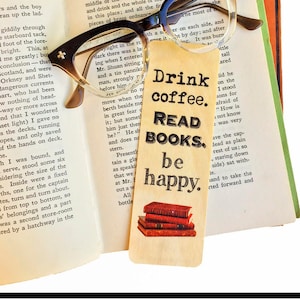 May include: A wooden bookmark with the text "Drink coffee. Read books. Be happy." and a stack of books on top. The bookmark is sitting on top of an open book with a pair of glasses on top of the book.