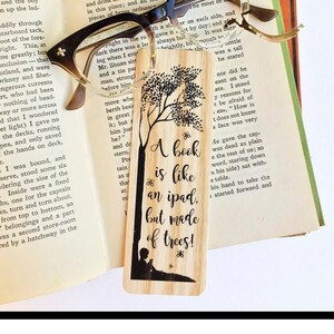 May include: A wooden bookmark with a black and white illustration of a tree and a person reading a book. The bookmark has the text "A book is like an ipad, but made of trees!"