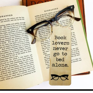 May include: Wooden bookmark with a brown background and black text that reads "Book lovers never go to bed alone."  The bookmark features a pair of black glasses with a brown frame.