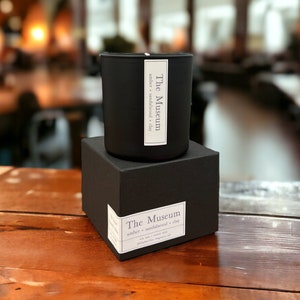 Op de afbeelding: Een zwarte kaars pot met een wit label waarop staat "The Museum amber + sandalwood + clay". De kaars staat op een zwarte doos met hetzelfde label.