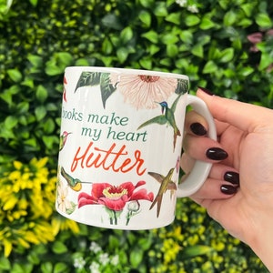 May include: White ceramic mug with a floral design and hummingbirds. The mug has the text "Books make my heart flutter" in red and orange.